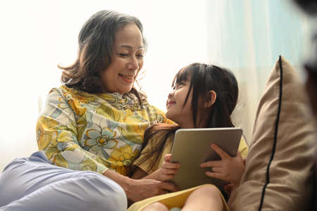 Happy middle aged woman embracing little preschool granddaughter, using digital tablet together on couch at home.の写真素材