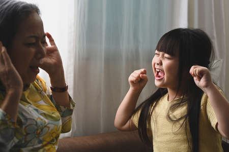 Unhappy grandmother having problem with noisy naughty little granddaughter screaming and covering ears. Naughty difficult rebellious child girl problems concept.の写真素材