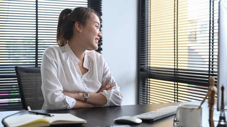 Smiling Asian woman entrepreneur sitting in her personal office and looking through the glass window.の写真素材