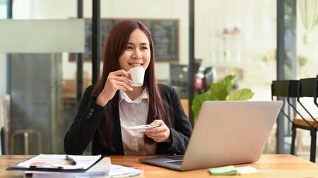 Thoughtful asian woman in business suit drinking hot coffee and using laptop at modern coffee shop.