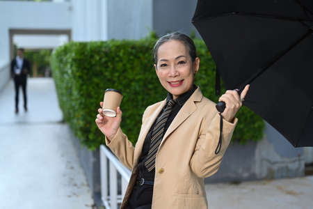 Smiling mature business woman holding takeaway coffee cup and sitting on bench under umbrella at city street.の写真素材