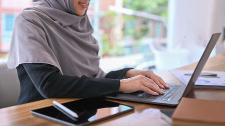 Smiling female muslim office worker using laptop computer while sitting at modern workplace.の写真素材