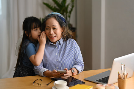 Adorable little girl whispering something to happy senior grandmother, spending leisure weekend at home together. Loving family relationship concept.の写真素材