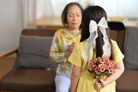Rear view of grandchild making surprise, standing with bouquet in hands and excited grandmother sit with eyes closed on sofa.の写真素材