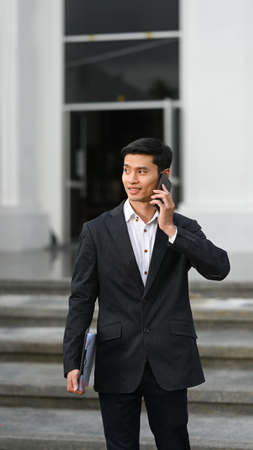 Handsome businessman in black suit standing near modern office building and talking on cell phone.の写真素材