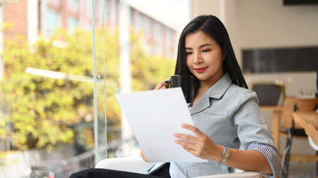 Attractive asian female manager sitting near large window in modern office and checking annual reports.の写真素材