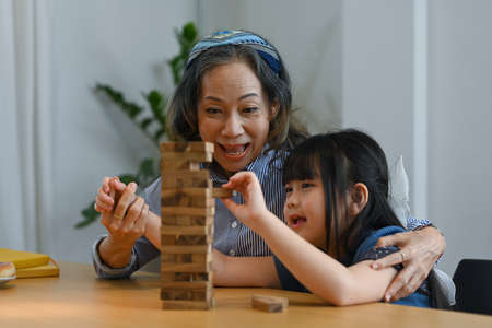 Little asian child and grandmother having fun playing wood block stacking board game together at home.の写真素材