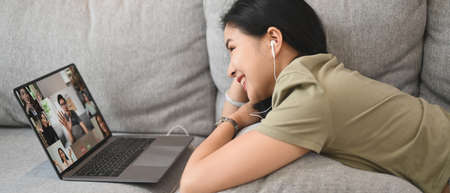 Cheerful young woman communicating by video conference via laptop while lying on couch at home.