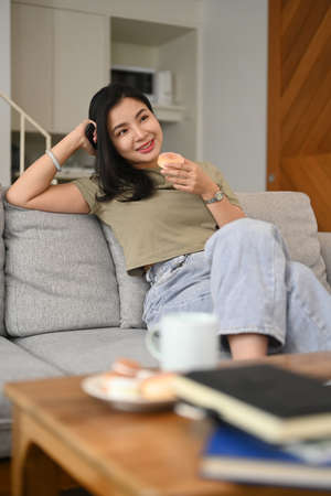 Smiling young woman eating donut and watching television on comfortable sofa at home.の写真素材