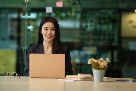 Confident female entrepreneur sitting in modern office with laptop computer and smiling at camera.の写真素材