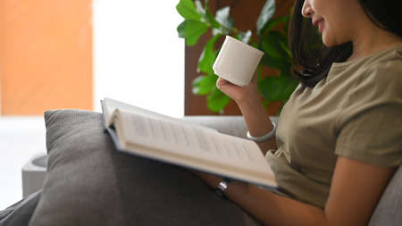 Calm asian woman drinking hot tea and reading book on comfortable couch at home.の写真素材