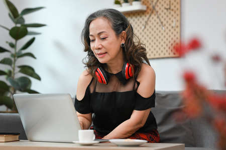 Smiling mature woman surfing internet, using bank service, sitting on cozy sofa in living room.の写真素材