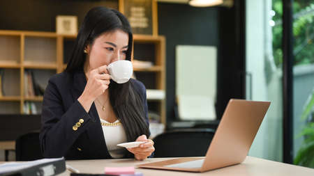 Attractive female entrepreneur drinking coffee an reading information online on laptop computer.の写真素材