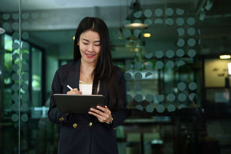 Asian woman manager writing some information on document while standing in corporate office.の写真素材