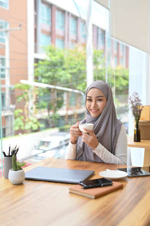 Smiling muslim businesswoman in hijab sitting in her office and enjoying morning coffee.の写真素材