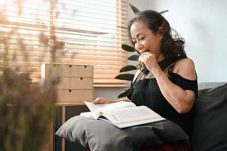 Peaceful middle aged woman eating cookie and reading book on couch at home. Leisure activity, positive mood concept.の写真素材