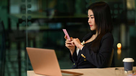 Pleasant asian businesswoman sitting in contemporary office and using mobile phone.