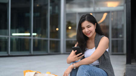 Smiling Asian woman sitting on stairs in front of shopping mall and using smart phone.