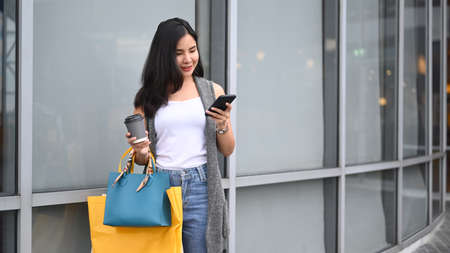 Pretty Asian woman using smart phone while walking down a street in the shopping district with her...