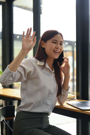 Cheerful female employee waving hands to greet her colleagues while talking on mobile phone.の写真素材