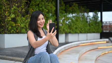 Cheerful asian woman having video call with friend on mobile phone while sitting in a park on a...
