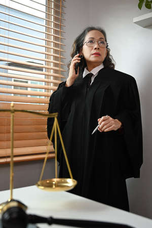 Experienced mature female lawyer standing in her office and talking on mobile phone with her clients for providing law consultation.の写真素材