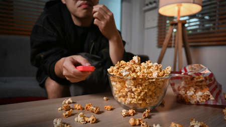 Joyful young asian man eating popcorn and watching TV at home in cozy living room.の写真素材