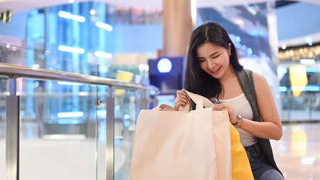 Happy young asian woman sitting on bench in shopping mall with paper bags.の写真素材