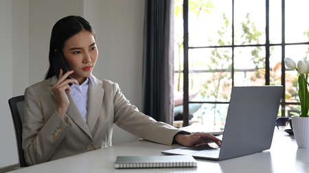 Serious female entrepreneur checking online information on laptop and talking on mobile phone.の写真素材