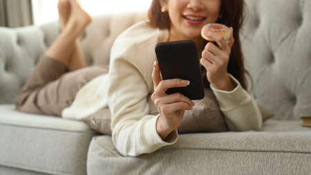 Smiling young woman in sweater lying on comfortable couch and chatting online via smart phone.の写真素材