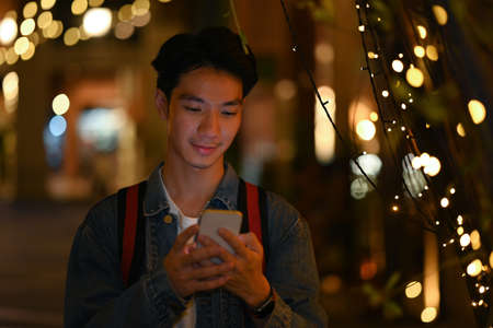 Smiling young asian male typing text message on smart phone while standing in the night city streets.の写真素材