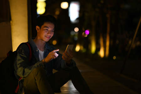 Man using smart phone while sitting on stairs in the night city with blurred night street lights background.の写真素材