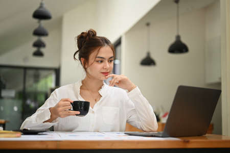 Gorgeous young businesswoman with cup of coffee in her hand reading information or business email on...