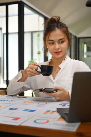 Shot of an attractive young businesswoman drinking coffee and using laptop in her workstation.の写真素材