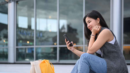Stylish young asian woman sitting on the stairs outdoors and using smart phone.