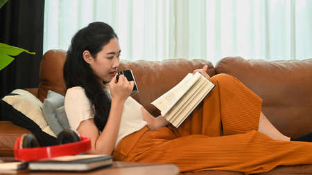 Peaceful young woman drinking hot tea and reading book on cozy couch at home.
