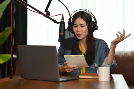 Smiling asian woman radio host using microphone and laptop recording podcast in small house studio.の写真素材