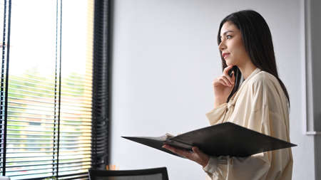 Pensive female office worker standing in modern office and looking out of window, dreaming about...