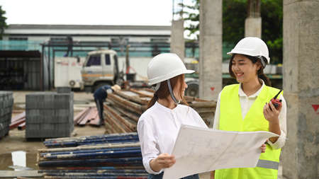 Smiling female real estate investor and civil engineers manager wearing safety helmets examining plans at construction site.の写真素材