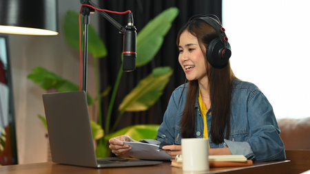 Woman podcaster in headphone using laptop and talking into a microphone while streaming her podcast...