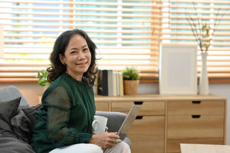 Satisfied middle aged woman sitting in bright living room and using laptop. Retirement, enjoy life...