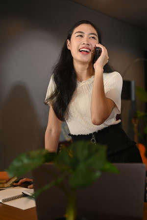 Young female entrepreneur having pleasant phone conversation with her business partner, sitting in home office.の写真素材