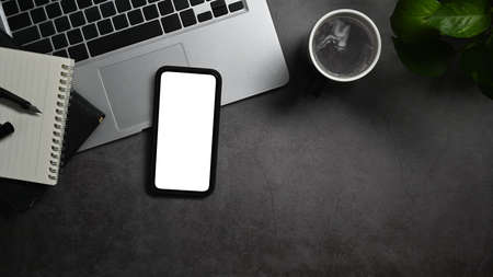 Smart phone with white screen, laptop, notebook and coffee cup on black stone table. Top view, flat...
