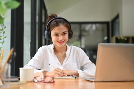 Attractive female student wearing headphone, listening lecture during study online on laptop. Education, e-learning, distance training.の写真素材
