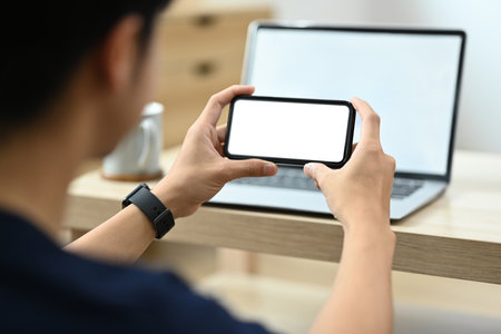 Over shoulder view of man sitting front of laptop and using mobile phone. Blank screen for your advertise text.の写真素材