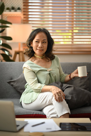 Beautiful middle aged woman relaxing on couch, enjoying stress free peaceful mood wellbeing in cozy living room.の写真素材