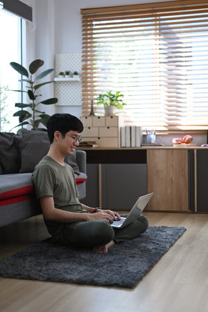 Asian man in casual clothes using laptop while sitting on carpet in cozy living room.の写真素材