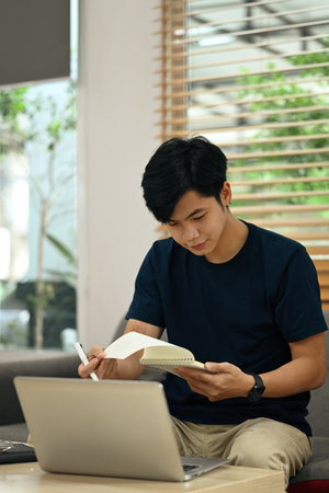 Focused asian male freelancer working online on laptop and making notes on notepad.の写真素材