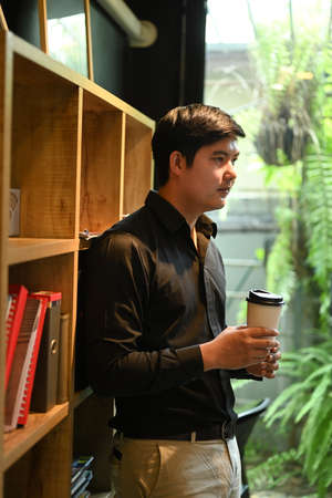 Portrait of asian businessman holding paper cup of coffee standing near bookshelves in his personal office.の写真素材