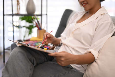 Relaxed young woman sitting on couch and painting picture with watercolor. Art, creative hobby and...
